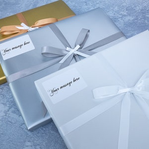 May include: Three gift boxes wrapped in gold, silver, and white paper with white satin ribbon bows. Each box has a white label with the text "Your message here".