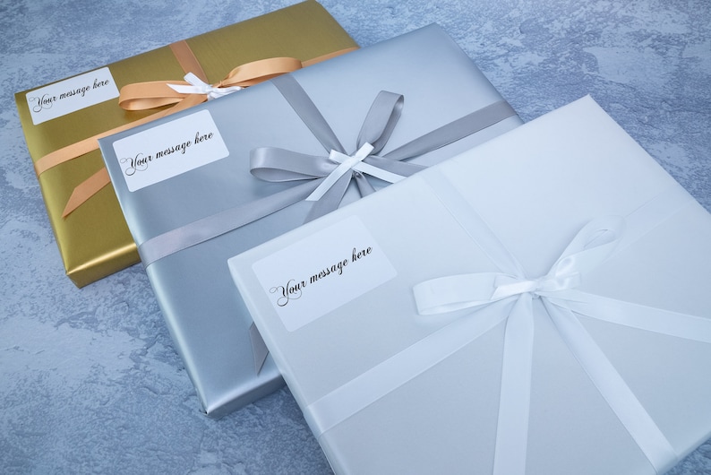 May include: Three gift boxes wrapped in gold, silver, and white paper with white satin ribbon bows. Each box has a white label with the text "Your message here".
