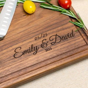 May include: A personalized walnut wood cutting board with the text "Emily & David 05/25 2023" engraved on the surface. The board has a natural wood grain and a dark brown color.