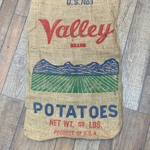 May include: A burlap potato sack with the words "U.S. No 1 Valley Brand Potatoes" printed in red and blue. The sack features a graphic of a green field and blue mountains. The net weight is 50 lbs.