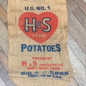 May include: A burlap potato sack with the text "U.S. NO. 1" and "H&S BRAND POTATOES" in red and blue. The sack is labeled "50 LBS. - NET WT. - 22.68 KILOS" and "PRODUCE OF U.S.A."