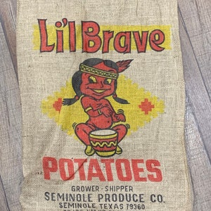 Peut inclure: Un sac en toile de jute vintage pour les pommes de terre avec une illustration de dessin animé d'un Amérindien jouant du tambour. Le sac est étiqueté "Li'l Brave Potatoes" et "Seminole Produce Co."