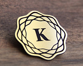 May include: A gold-coloured metal badge with a black outline and a black letter "K" in the centre.
