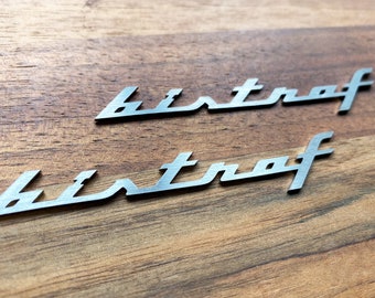 May include: Two silver-colored "bistraf" wordmark emblems are displayed on a wooden surface. The wordmark has a stylized, cursive font. The wood grain is visible, adding a natural texture to the background.