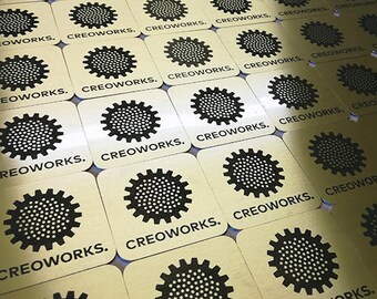 May include: A close-up of a large quantity of gold-coloured metal coasters with a black gear design and the text "CREOWORKS." printed on each coaster.