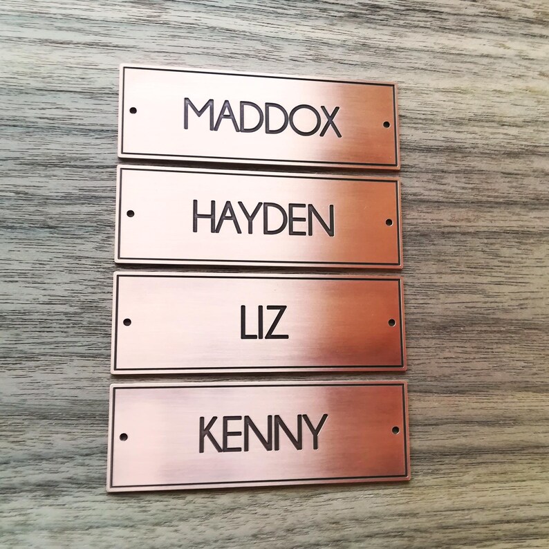 May include: Four rectangular copper-colored nameplates with black lettering. The names on the plates are Maddox, Hayden, Liz, and Kenny.