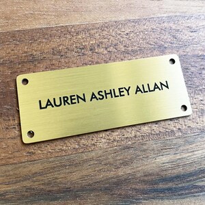 Peut inclure: Une plaque de nom en métal doré avec le texte "LAUREN ASHLEY ALLAN" gravé en noir.