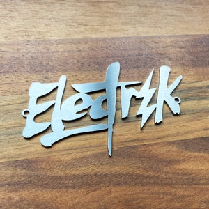 Op de afbeelding: Een zilveren metalen bord met het woord "Elektrik" geschreven in een gestileerd, graffiti-achtig lettertype.