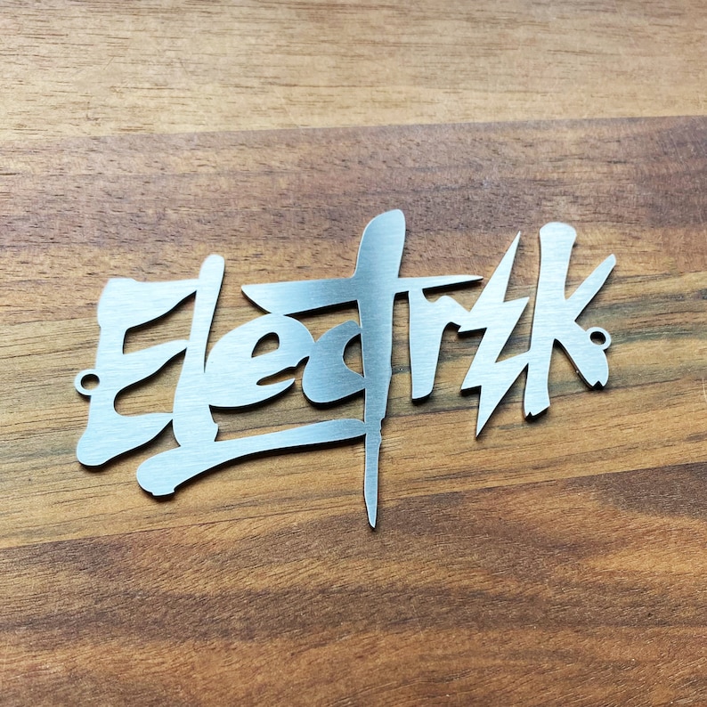 Op de afbeelding: Een zilveren metalen bord met het woord "Elektrik" geschreven in een gestileerd, graffiti-achtig lettertype.