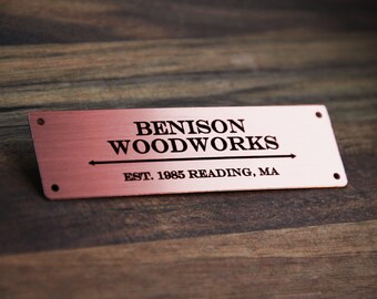 May include: A copper-coloured metal sign with black engraved text that reads "Benison Woodworks Est. 1985 Reading, MA". The sign has a brushed metal finish and a subtle arrow pointing to the right.