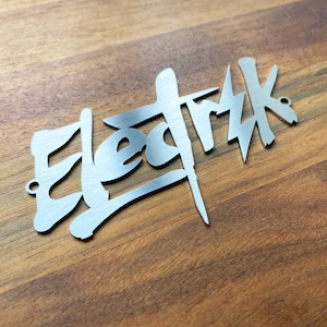 Op de afbeelding: Een zilveren metalen bord met het woord "Elektrik" geschreven in een gestileerd, graffiti-achtig lettertype.
