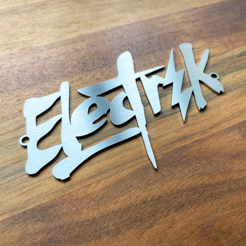 Op de afbeelding: Een zilveren metalen bord met het woord "Elektrik" geschreven in een gestileerd, graffiti-achtig lettertype.