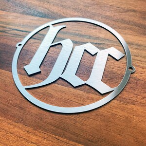 Op de afbeelding: Een zilveren metalen cirkel met de letters "hcc" in een gotisch lettertype.