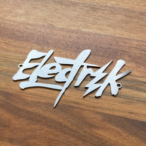 Op de afbeelding: Een zilveren metalen bord met het woord "Elektrik" geschreven in een gestileerd graffiti-lettertype.