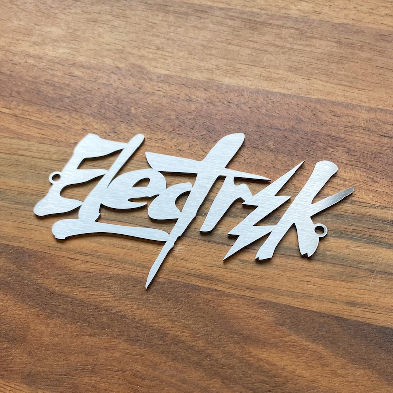 Op de afbeelding: Een zilveren metalen bord met het woord "Elektrik" geschreven in een gestileerd graffiti-lettertype.