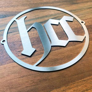 Op de afbeelding: Een zilveren metalen cirkel met het woord "HOC" in een gotisch lettertype.