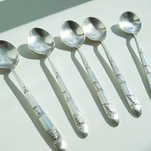 May include: Six silver spoons with oval bowls and long, decorated handles. The spoons are arranged diagonally, casting shadows on a white surface. The handles feature geometric patterns and are slightly tapered.