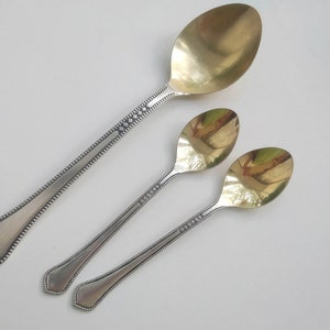 May include: Three spoons with silver handles and gold-colored bowls are arranged diagonally on a white surface. The handles have a decorative pattern of small diamonds and dots. The largest spoon is at the top, with two smaller spoons below.