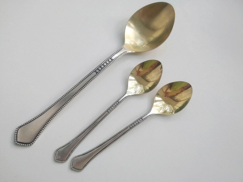 May include: Three spoons with silver handles and gold-colored bowls are arranged diagonally on a white surface. The handles have a decorative pattern of small diamonds and dots. The largest spoon is at the top, with two smaller spoons below.