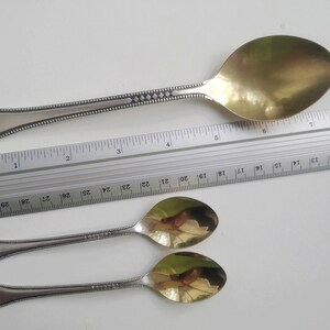 May include: A large silver serving spoon with a gold-colored bowl, approximately 10 inches long, and two smaller silver spoons. The spoons have decorative handles with a beaded and diamond pattern. A ruler is placed next to the spoons.
