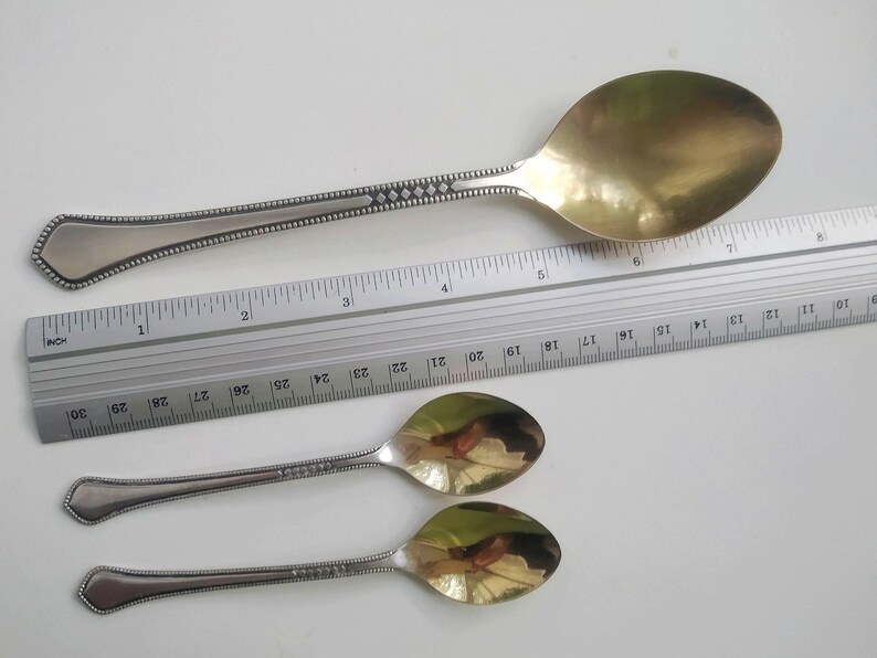May include: A large silver serving spoon with a gold-colored bowl, approximately 10 inches long, and two smaller silver spoons. The spoons have decorative handles with a beaded and diamond pattern. A ruler is placed next to the spoons.