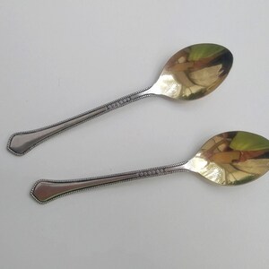 May include: Two antique silver spoons with gold-colored bowls. The handles are detailed with a dotted pattern. The spoons are displayed on a white surface, reflecting the light.