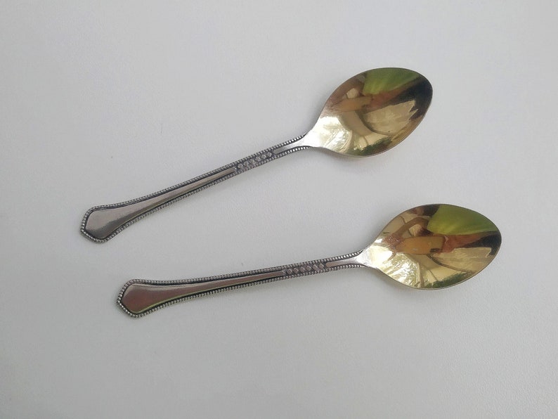 May include: Two antique silver spoons with gold-colored bowls. The handles are detailed with a dotted pattern. The spoons are displayed on a white surface, reflecting the light.