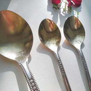 May include: Three silver spoons of varying sizes are arranged on a white surface. The spoons have oval bowls and decorative handles with a repeating diamond and dot pattern. The spoons reflect the light, creating a bright, shiny appearance.