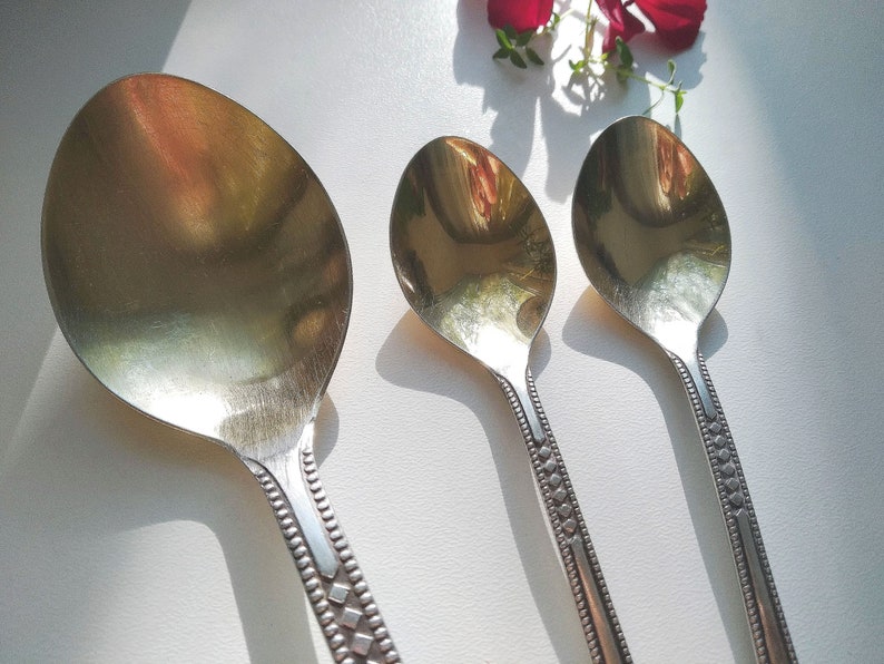 May include: Three silver spoons of varying sizes are arranged on a white surface. The spoons have oval bowls and decorative handles with a repeating diamond and dot pattern. The spoons reflect the light, creating a bright, shiny appearance.