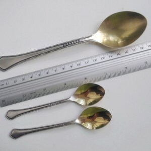 May include: A large silver serving spoon with a gold-colored bowl, and two smaller spoons. The spoons have decorative detailing on the handles. A ruler is placed next to the spoons for scale.