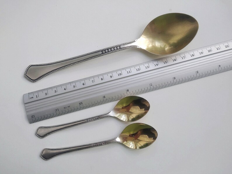 May include: A large silver serving spoon with a gold-colored bowl, and two smaller spoons. The spoons have decorative detailing on the handles. A ruler is placed next to the spoons for scale.