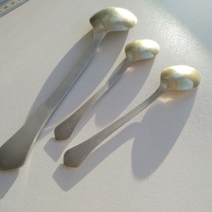 May include: Four silver spoons of varying sizes are arranged on a white surface. The spoons have elongated handles and rounded bowls. The spoons are made of a shiny, reflective metal, and cast shadows.