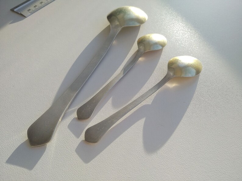 May include: Four silver spoons of varying sizes are arranged on a white surface. The spoons have elongated handles and rounded bowls. The spoons are made of a shiny, reflective metal, and cast shadows.