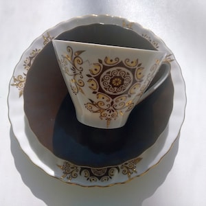 May include: A white teacup, saucer, and plate set with gold and brown floral designs. The teacup is centered on the saucer, which rests on a larger plate. The edges of the plates and cup are scalloped and trimmed with gold.