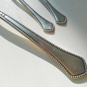 May include: Close-up of three silver spoons with a decorative beaded edge and diamond-shaped accents on the handle. The spoons are arranged on a white surface, with sunlight casting shadows.