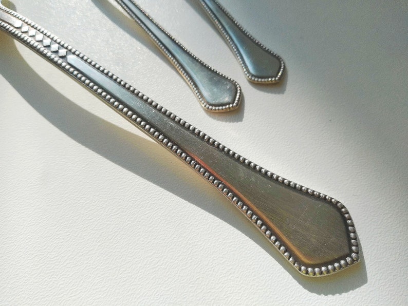 May include: Close-up of three silver spoons with a decorative beaded edge and diamond-shaped accents on the handle. The spoons are arranged on a white surface, with sunlight casting shadows.