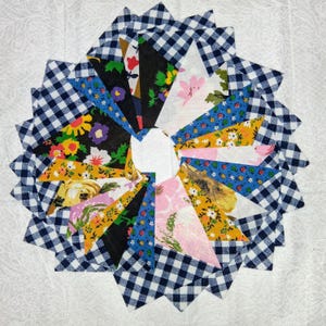 May include: A circular patchwork design featuring a central white circle surrounded by alternating layers of blue and white gingham and colorful floral fabric triangles. The floral fabrics include yellow, pink, blue, and black patterns.