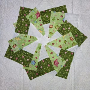 May include: A handmade quilt block featuring a starburst design. The block is composed of green fabric with floral and star patterns, and white fabric. The star-shaped design is centered on a white background.