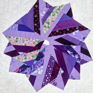 May include: A star-shaped quilt block composed of various fabric strips in shades of purple, lavender, and violet. The design features a mix of solid colors, floral patterns, star prints, and galaxy motifs arranged in a radiating, geometric pattern.