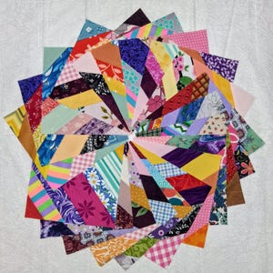 May include: A circular arrangement of fabric scraps featuring a diverse array of colors and patterns, including floral, plaid, polka dot, striped, and geometric designs. The pieces are layered in a sunburst pattern, creating a vibrant, multi-textured display.