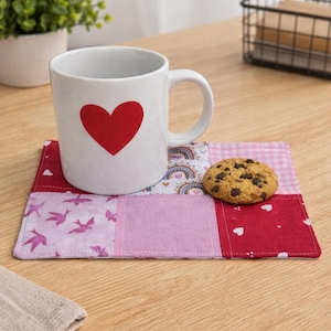 May include: A white ceramic mug with a red heart design sits on a patchwork coaster. The coaster features various pink and red fabrics with bird and heart patterns. A chocolate chip cookie rests on the coaster, creating a cozy, inviting scene.