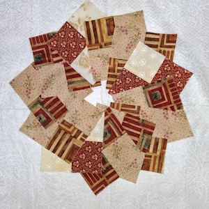 May include: A patchwork quilt block with a starburst design. The block is composed of various fabric squares in shades of beige, cream, and red. Some squares feature floral patterns, while others have geometric designs. The quilt block is set against a white background.