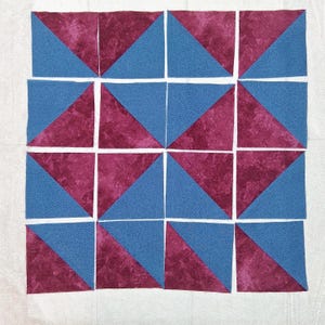 May include: Sixteen fabric squares arranged in a grid pattern. Each square is divided diagonally, featuring a blue triangle and a burgundy triangle. The fabric has a textured appearance, and the squares are separated by white seams. The overall design suggests a quilting project.
