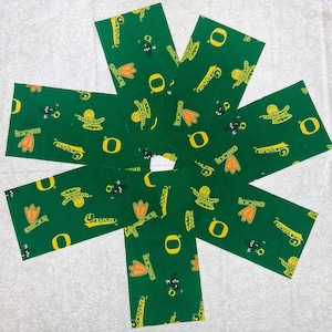 May include: A set of six rectangular fabric strips in dark green, featuring a repeating pattern of yellow Oregon Ducks logos, the word Oregon, and orange duck feet. The fabric pieces are arranged in a circular, fan-like formation.
