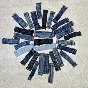 May include: A collection of rectangular denim fabric strips in various shades of blue, black, and light blue, arranged in a circular pattern. Each strip features visible stitching along the edges, showcasing different denim textures and washes.