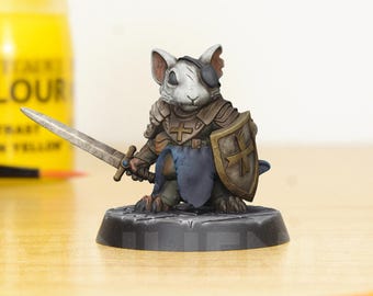 May include: A miniature figurine of a white mouse wearing armor and holding a sword and shield. The mouse is wearing a blue cape and has a white cross on its chest. The figurine is standing on a black base.