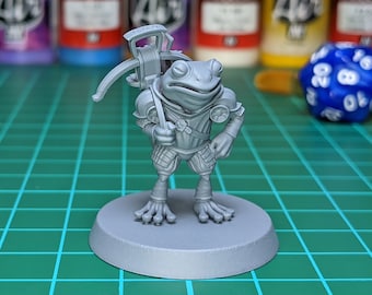 May include: A gray, unpainted miniature figurine of a frog wearing armor and holding a crossbow. The frog is standing on a round base. The figurine is on a green surface.