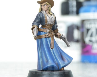 Op de afbeelding: Miniatuurfiguur van een vrouwelijke revolverheld. Ze draagt een bruine cowboyhoed, een blauwe jurk en een bruin korset met bijpassende riem. De figuur houdt twee zilveren revolvers vast. De basis is donkergrijs. Gedetailleerd geschilderd.
