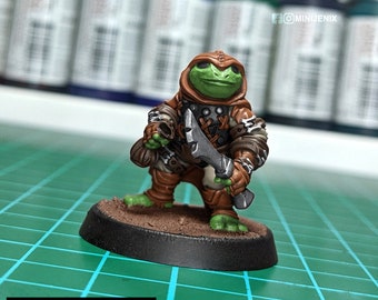 May include: A green and brown painted miniature figurine of a frog-like creature wearing a hooded cloak and holding a dagger. The figurine is standing on a round brown base.