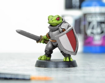 May include: A green frog miniature figure wearing silver armor and holding a sword and a shield with a red and white design. The miniature is on a black base.